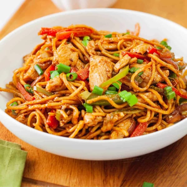 A white bowl filled with chicken noodles, red and green bell peppers, and green onions.