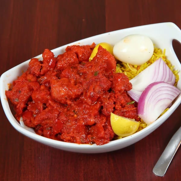 Indian chicken tikka in a white bowl with yellow rice, lemon wedges, red onion, and a boiled egg.
